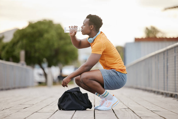 Afroamerikkalainen mies juo vettä sillalla. Mies pitää taukoa pullosta juomisesta. Urheileva kaveri keskittyy terveyteen. - Valokuva, kuva