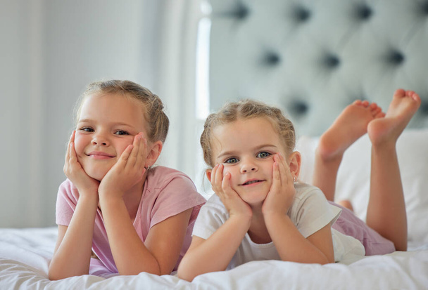 Portrait of two little girls lying on a bed at home. Smiling girls only relaxing and looking comfy, two blonde caucasian sisters enjoy time in bed together in a bright bedroom at home. - Foto, afbeelding