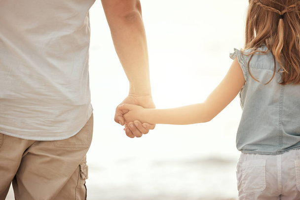 Egy apa és lánya kézen fogva a parton naplementekor. Apa kötődik a gyerekéhez, gyengéd, és vigyáz rá azzal, hogy kint tartja a kezét. Szülő és gyermek együtt. - Fotó, kép