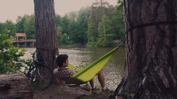 Man traveling by bicycle, resting in green hammock in woods by lake. Cyclist in hammock at campsite by river. Man with bike in hammock looking out into distance. Active leisure and sports activities - Filmati, video