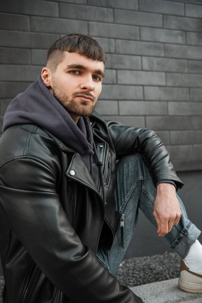 Fashion hipster handsome man with tattoos on neck in casual clothes with leather jacket, hoodie and jeans sits and poses near a white and brick brick wall on the street - Photo, Image