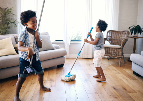 İki melez küçük çocuk evdeki salonda süpürge ve paspasla oynuyorlar. Mutlu kardeşler evde temizlik malzemeleriyle oynarken eğleniyorlar. Evde temizlik yaparken kaygısız kardeşler şarkı söylüyor.. - Fotoğraf, Görsel