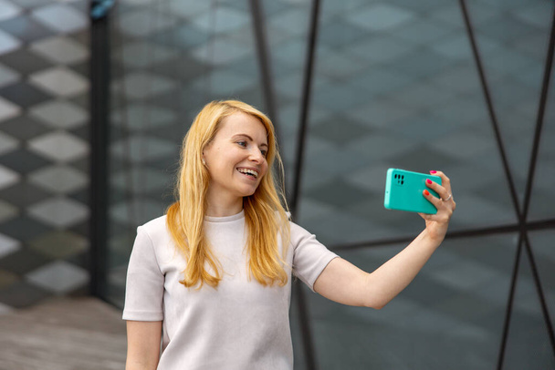 Μια νεαρή ξανθιά γυναίκα βγάζει selfie στο smartphone για τους οπαδούς της. Νεαρή κοπέλα με επιρροή. Δημοτικότητα στο διαδίκτυο, φίλοι σε απευθείας σύνδεση. Όμορφο κορίτσι κάνει περιεχόμενο για λογαριασμό της στα μέσα κοινωνικής δικτύωσης - Φωτογραφία, εικόνα