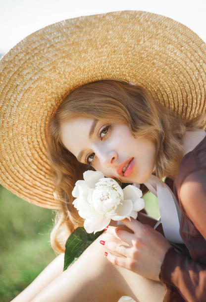 Portrait of a cute blonde girl in a straw hat posing with her arms around her knees, holding a peony flower in her hand. - Photo, image