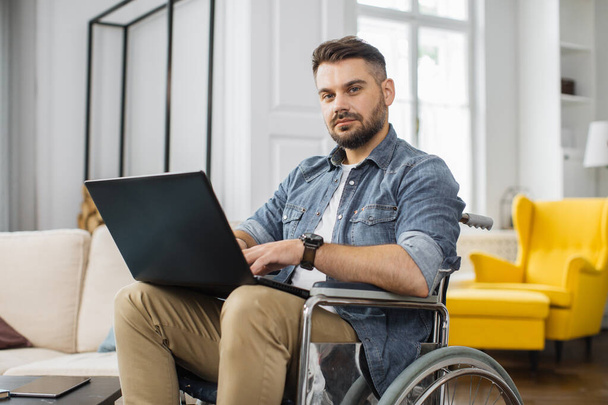 Handsome disabled man sitting in wheelchair with laptop on knees and looking at camera. Caucasian male in casual wear having opportunities to remote work at home. - Photo, Image