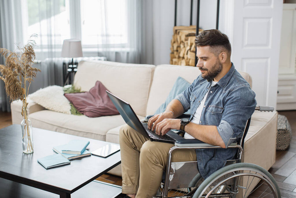 Side view of concentrated young man in wheelchair typing on wireless laptop in bright living room. Caucasian male employee with disability working at international company from home. - Photo, image