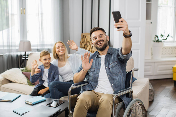 Caucasian man with disabilities holding smartphone and taking selfie with family at home. Smiling mother and son sitting together on sofa near father in wheelchair and waving hands on phone camera. - Foto, Imagem