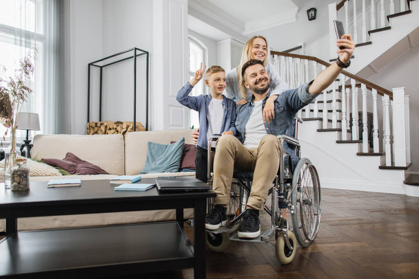 Young man sitting in wheelchair while shooting selfie on modern smartphone with wife and son standing near. Happy caucasian family smiling and looking at device camera in bright living room. - Valokuva, kuva
