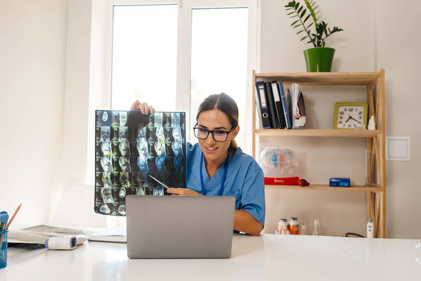 White woman doctor smiling while working with laptop and x-ray in office indoors - Foto, Imagen