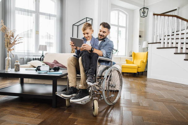 Smiling dad with special needs sitting in wheelchair and holding son on knees while playing on digital tablet. Positive family using portable device for playing childrens educational games. - 写真・画像