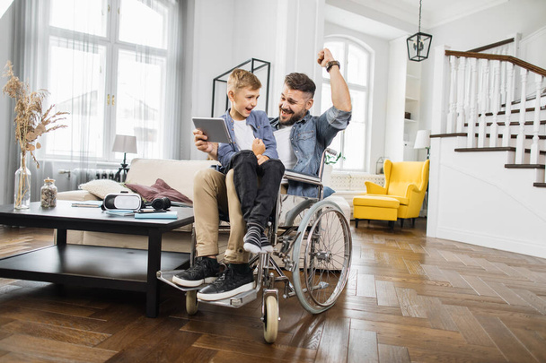 Emotional male wheelchair user enjoying video racing victory on modern tablet with little son. Family clenching fists and smiling while celebrating success in living room. - Fotografie, Obrázek