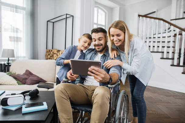 Happy dad in wheelchair holding modern tablet and showing funny video to standing nearby wife with son. Joyful family smiling while looking at device screen in trendy living room. - 写真・画像