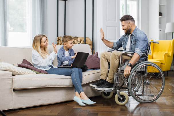 Bearded husband in wheelchair waving hello gesture to little son and attractive wife at home. Young caucasian mother and child using wireless laptop for education. - Фото, изображение