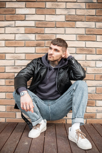 Stylish handsome hipster man in casual clothes with jacket, hoodies, jeans and sneakers sits near a brick wall in the city - Foto, Imagen