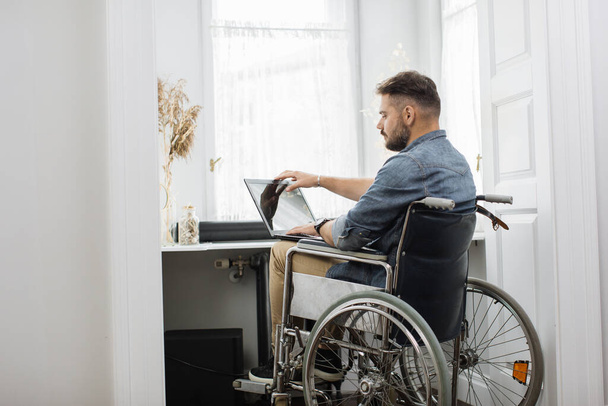 Handsome serious man in wheelchair finishing his remote work and closing modern laptop at home office. Concept of freelance, temporary disability and technology. - 写真・画像