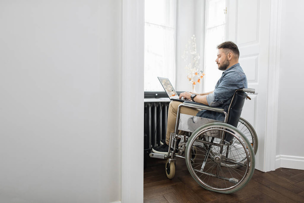 Mężczyzna freelancer z niepełnosprawnością pracuje na bezprzewodowym laptopie w jasnym pokoju w domu. Brodaty mężczyzna w luźnych ubraniach siedzi na wózku inwalidzkim w pobliżu dużego okna. - Zdjęcie, obraz
