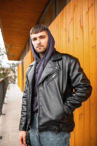 Stylish hipster street guy in a fashion black leather jacket and hoodie standing next to a wooden yellow wall - Fotó, kép