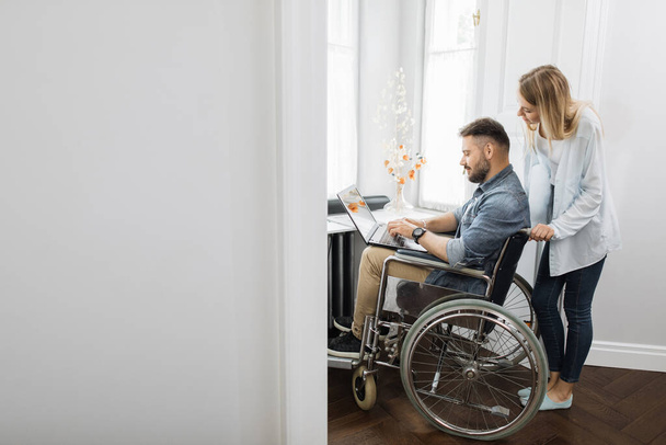 Disabled caucasian man sitting in wheelchair and working on modern laptop while his charming wife standing behind. Family support, people and love. - Foto, Imagem