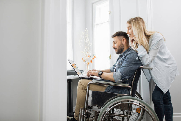 Bearded young man in wheelchair looking at laptop screen while beautiful wife standing behind at home office. Happy wife supporting beloved disabled husband. - Fotoğraf, Görsel
