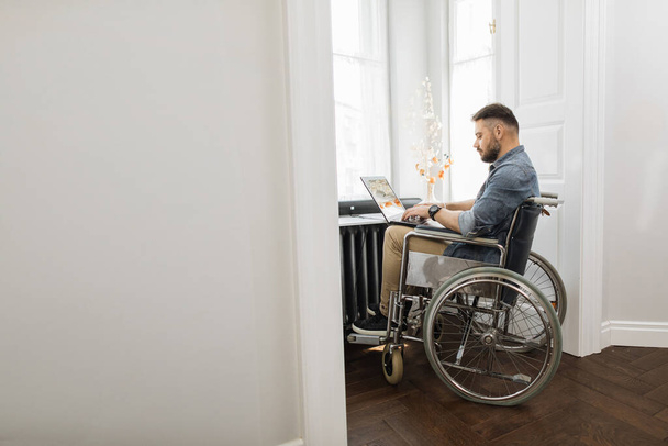Serious disabled young man using laptop for remote job at home. Confident caucasian male in wheelchair surfing internet near window at workplace. - 写真・画像