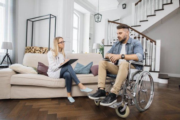 Young female doctor consulting disabled male patient and writing on clipboard at home. Rehabilitation specialist making medical checkup to man in wheelchair while sitting on sofa. - Photo, image
