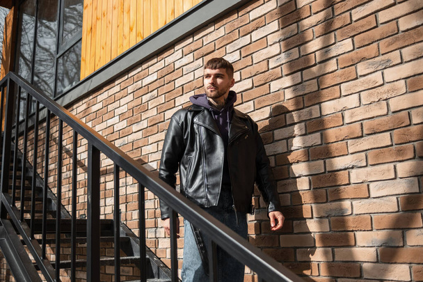 Cool brutal successful man hipster model with hair in fashionable clothes with a jacket and hoodie descends the metal staircase near the brick wall - Photo, Image