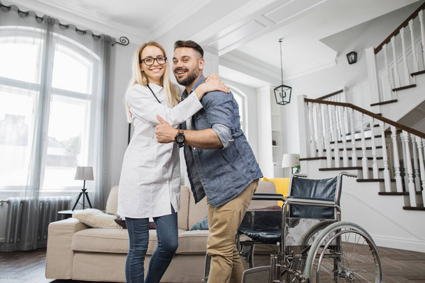 Portrait of positive female physiotherapist and bearded man during rehabilitation at home. Attractive woman embracing disabled man while smiling and looking at camera. - Foto, Bild
