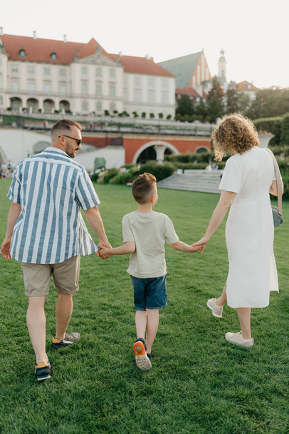 A photo from the back of a family is strolling the garden of the palace in an old European town. A happy father, mother, and son are holding hands at sunset. - Foto, imagen
