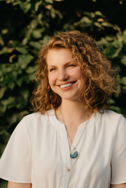 A smiling woman with curly hair is posing in the park at the sunset. A close portrait of a lady in a white dress with green leaves in the background. - Photo, Image