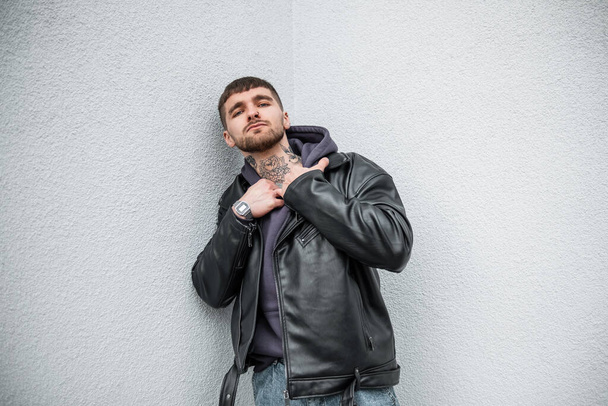 Fashionable handsome stylish hipster man with a tattoo on his neck in fashionable casual clothes with a leather rock jacket and hoodie stands and poses near a white wall on the street - Fotoğraf, Görsel