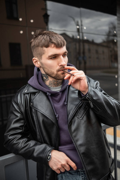 Жестокий красивый крутой модный мужчина в модной повседневной одежде в куртке и толстовке, курящий сигарету возле черного здания в городе - Фото, изображение