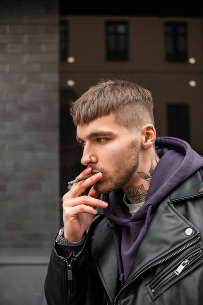 Street male portrait of a handsome brutal hipster man with a tattoo on his neck in fashionable casual clothing with a leather rock jacket and hoodie smoking a cigarette and walking in the city - Photo, Image
