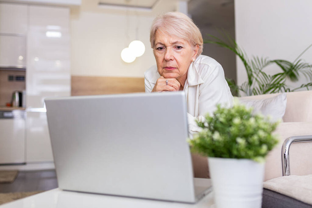 自宅のテーブルに座ってノートパソコンで作業している年配の女性の肖像画。ソファに座っている間に家からネットをサーフィンし、ラップトップコンピュータを使用している老婦人 - 写真・画像