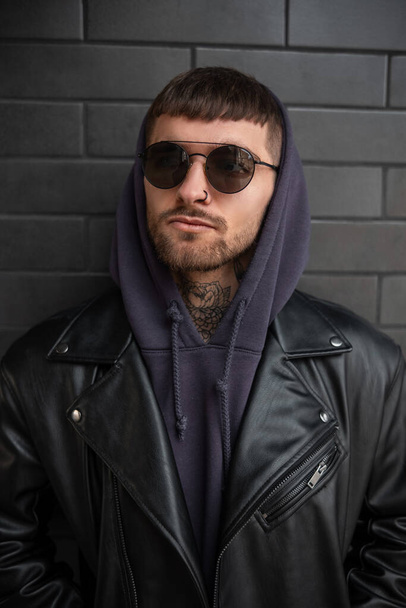 Men's street portrait of a handsome stylish hipster guy with round sunglasses in a trendy black leather jacket and hoodie near a black brick wall - Fotografie, Obrázek