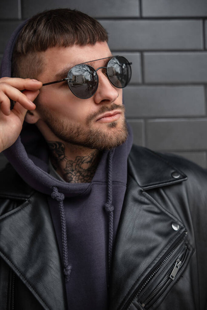 Retrato masculino urbano de hombre serio brutal guapo hipster en traje negro de moda con chaqueta de roca de cuero y sudadera con capucha lleva unas gafas de sol frescas sobre un fondo de ladrillo negro - Foto, imagen