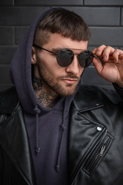 Fashion street male portrait of a handsome business stylish man in fashionable black sunglasses with a hood in a trendy black leather jacket and hoodie on a dark brick background on the street - Foto, Imagem