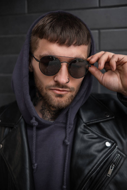 Fashionable hipster man model with hairstyle in stylish urban outfit with jacket and hoodie wears a fashion sunglasses near a brick black wall - Photo, Image