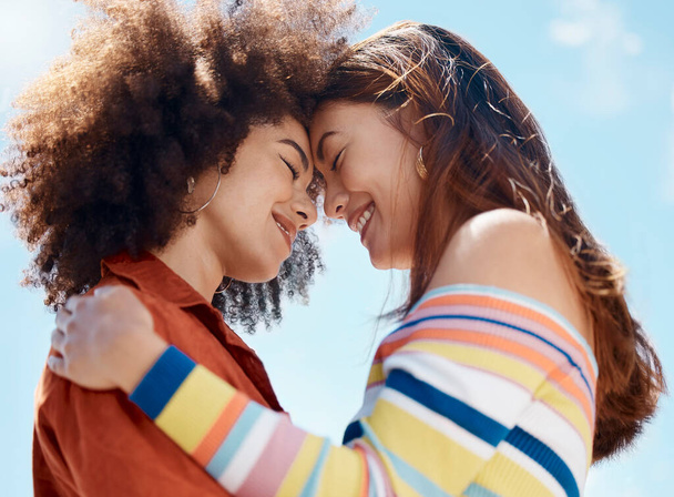 Close up of two young mixed race female couple embracing each other and smiling outside on a sunny day. A Beautiful gay hispanic woman with a cool afro hair style showing affection by hugging her - 写真・画像