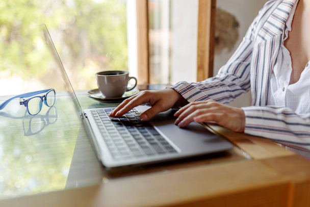 Обрізаний знімок молодої бізнес-леді, що працює на ноутбуці, сидячи в кафе або будинку, поруч з вікном
. - Фото, зображення
