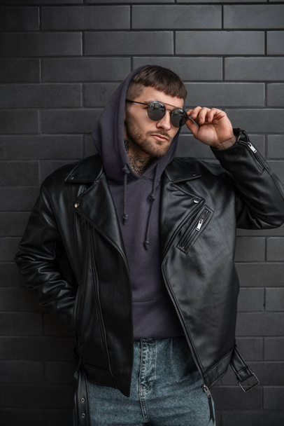Modeporträt eines gutaussehenden Mannes Hipster mit cooler stylischer Sonnenbrille in trendiger Lederjacke mit Kapuzenpulli posiert in der Nähe eines schwarzen modernen Backsteinhintergrundes - Foto, Bild