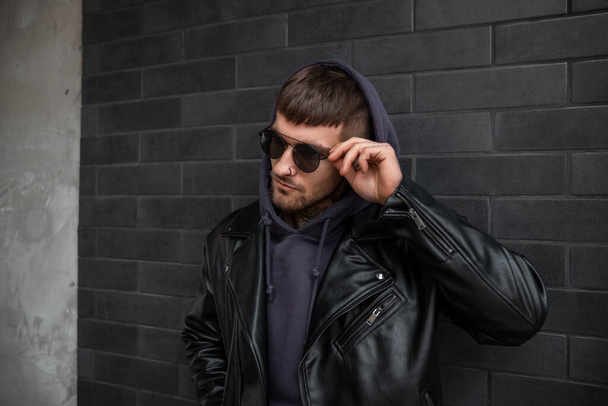 Modny przystojny facet w stylowych czarnych ubraniach w skórzanej kurtce i kapturze naprawia okulary przeciwsłoneczne i stoi przy ścianie z czarnej cegły na ulicy - Zdjęcie, obraz