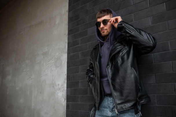 Cool handsome man model in fashionable clothes with leather rock jacket and hoodie is wearing stylish vintage sunglasses and standing near a black brick wall on the street - Photo, image