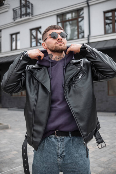 Cool stylish man model in a street fashion outfit with leather jacket and hoodie walking in the city - Фото, зображення