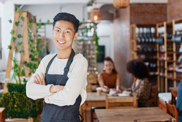 Portrait of one smiling handsome asian business owner standing with his arms crossed in his cafe. Confident and successful mixed race man wearing an apron in his restaurant. Ambitious entrepreneur. - Fotó, kép