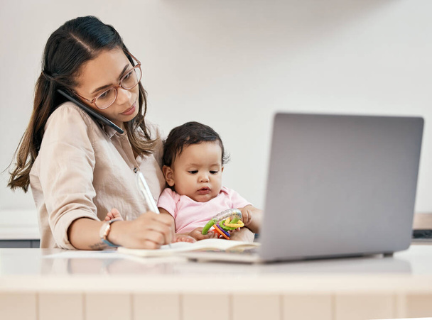 Shot of a woman working on her laptop and talking on her cellphone while sitting with her baby on her lap. - Foto, imagen