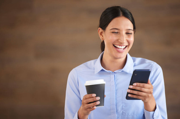 Smiling mixed race businesswoman isolated against a brown background with copyspace and using a cellphone to browse the internet during a coffee break. Hispanic professional laughing at social media. - 写真・画像