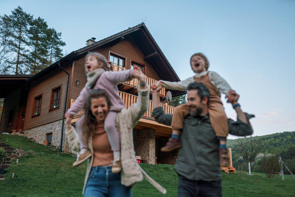 Happy parents with small children on piggybacks running and having fun together in a garden near their house. - Zdjęcie, obraz