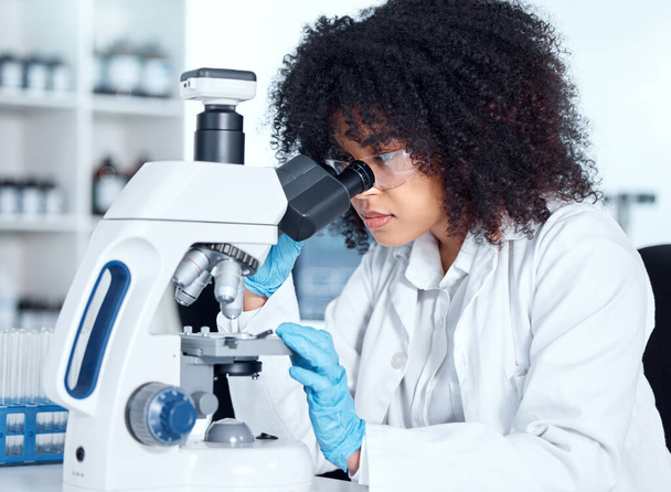 Young african american woman wearing a labcoat and goggles looking at medical samples on a microscope in her lab. A mixed race female scientist wearing goggles and gloves conducting research. - Fotó, kép