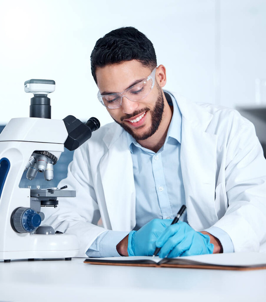 One handsome young mixed race man wearing gloves and a labcoat and looking at medical samples on a microscope in a lab. A male scientist wearing safety goggles and smiling while writing in a notebook. - Photo, Image
