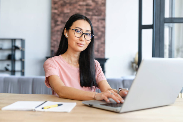 Elegant smart pretty Asian student or entrepreneur in glasses and earphones talking to business partner on the video call and typing, taking notes, and attentively listening and looking at the camera - Valokuva, kuva